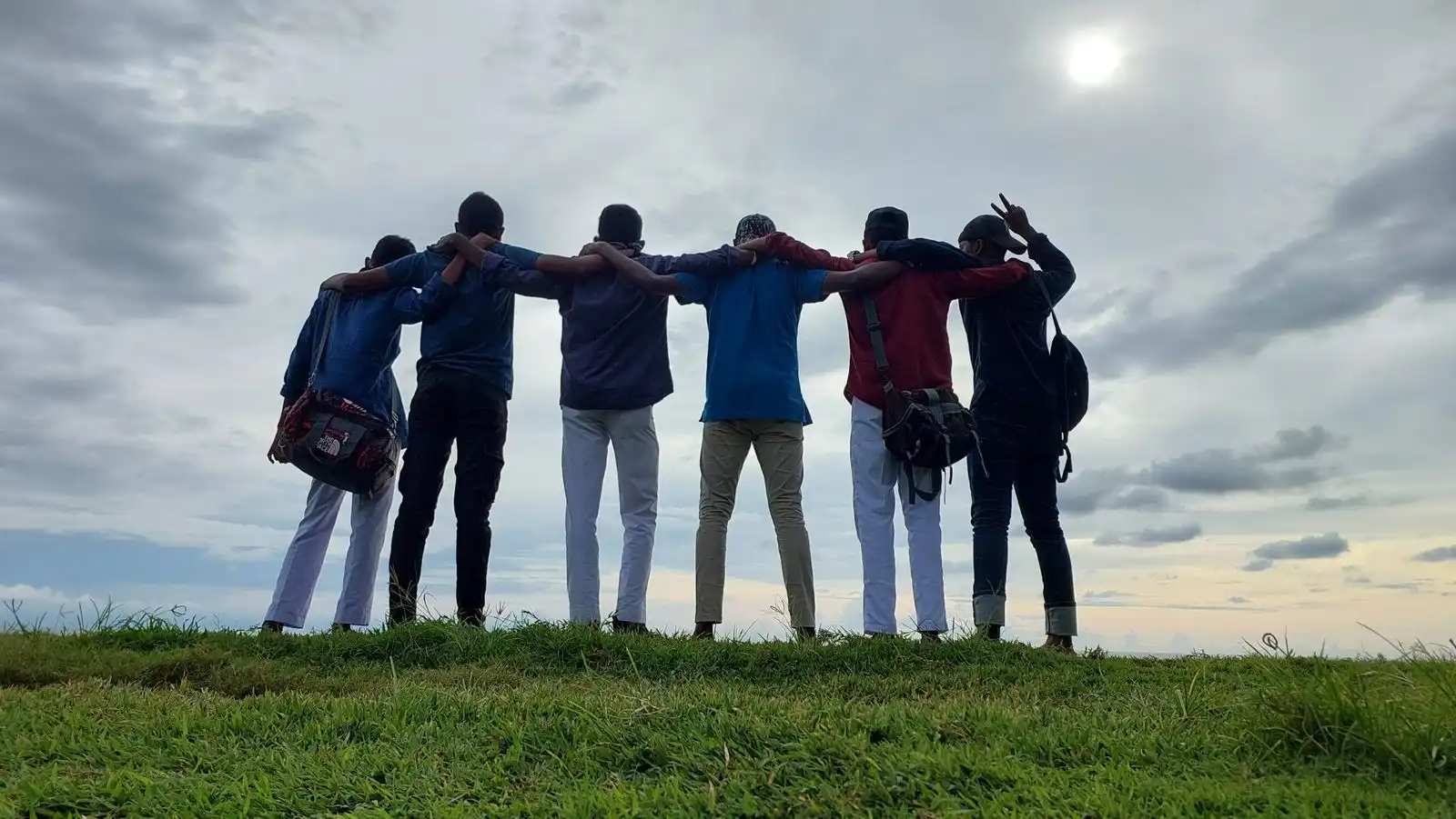 A group of friends standing arm in arm on a grassy hilltop under a cloudy sky, seen from behind