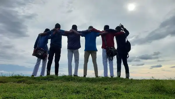 A group of friends standing arm in arm on a grassy hilltop under a cloudy sky, seen from behind