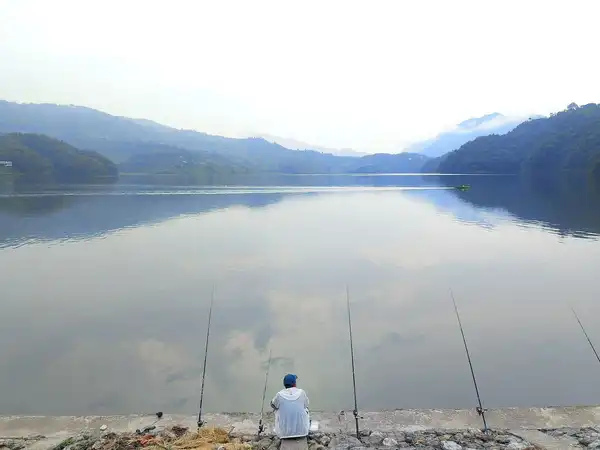 A still lake in morning mist below soft hills, with a lone figure fishing from the stony shore