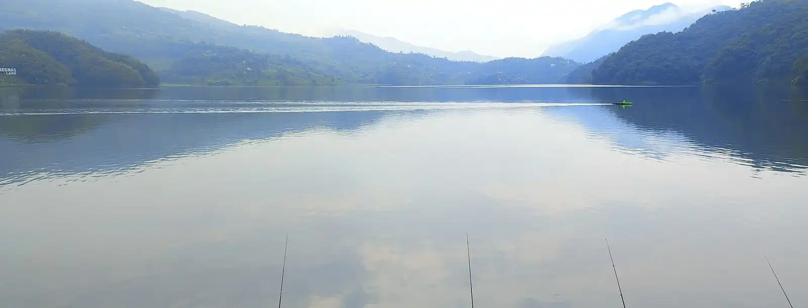 A still lake in morning mist below soft hills, with a lone figure fishing from the stony shore