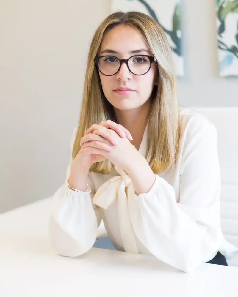 A woman with glasses and a cream blouse sitting at a white desk, hands clasped, looking at the camera