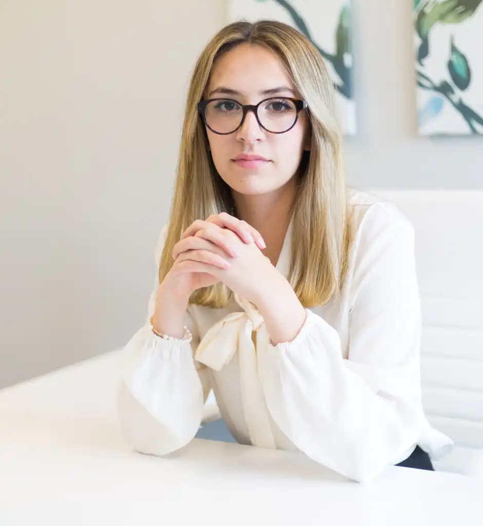 A woman with glasses and a cream blouse sitting at a white desk, hands clasped, looking at the camera