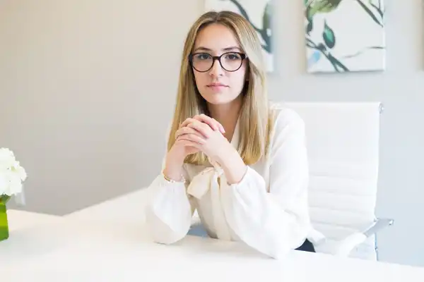 A woman with glasses and a cream blouse sitting at a white desk, hands clasped, looking at the camera