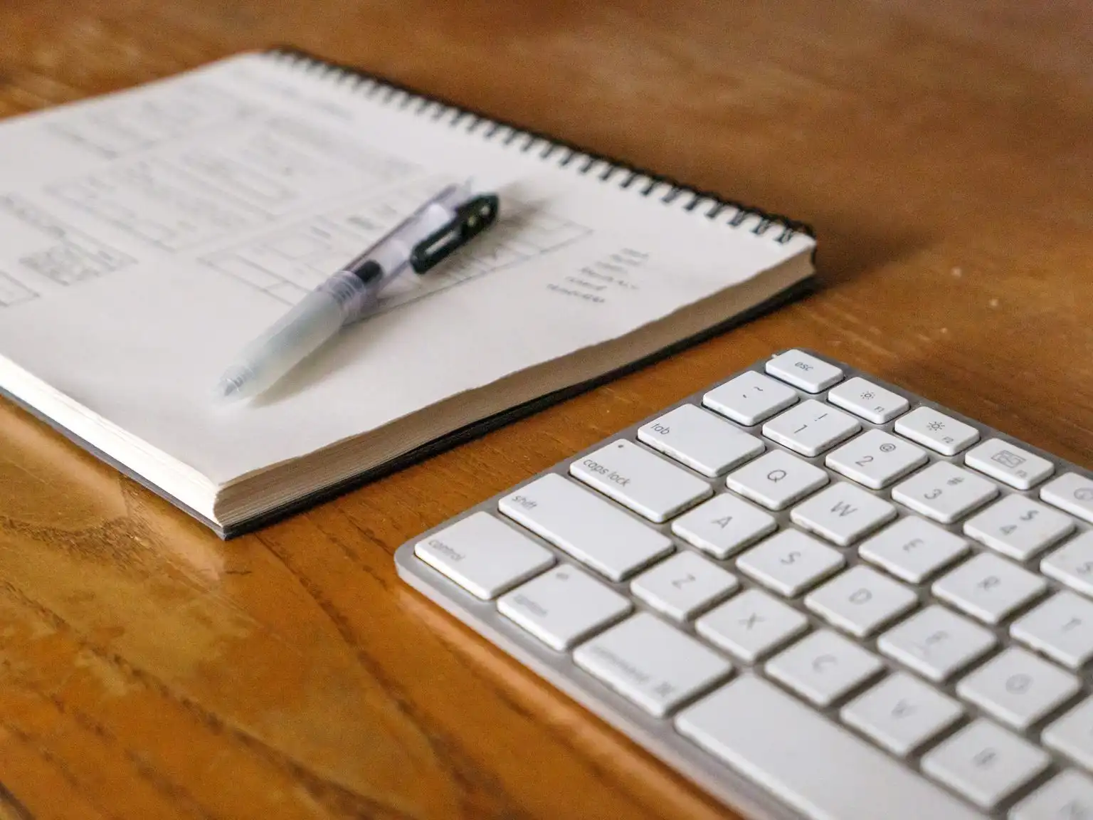 An open spiral notebook and a pen beside a white keyboard on a wooden desk